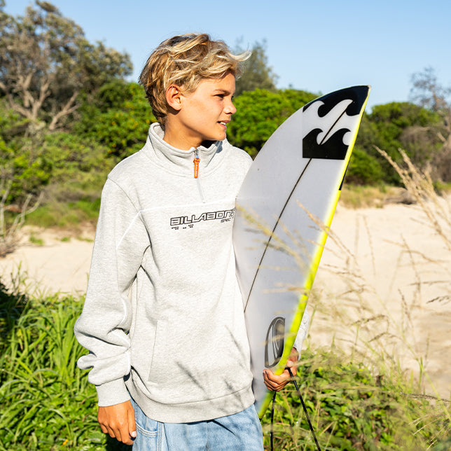 A boy wearing a light gray Billabong pullover stands outdoors holding a white surfboard with black graphics, with sandy dunes and green bushes in the background.