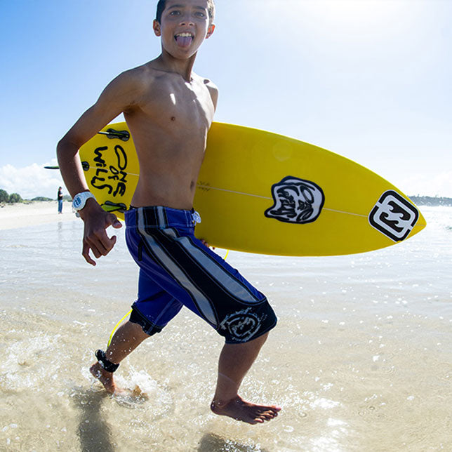 A teen boy running into shallow ocean water on a beach, holding a bright yellow surfboard under his arm. He is wearing blue and black board shorts, and the surfboard has black and white graphics.