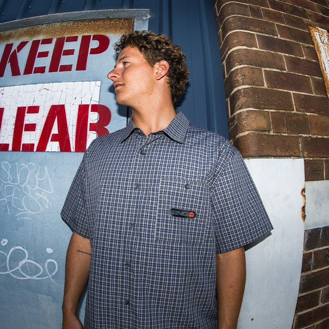 A man wearing a short-sleeve button-up shirt with a blue and white grid pattern, standing in front of a brick wall and a blue wall with red lettering.