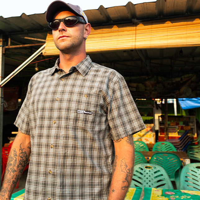 Man wearing sunglasses and a gray plaid shirt stands in front of colorful plastic chairs and a market stall. The sunny setting conveys a relaxed mood.