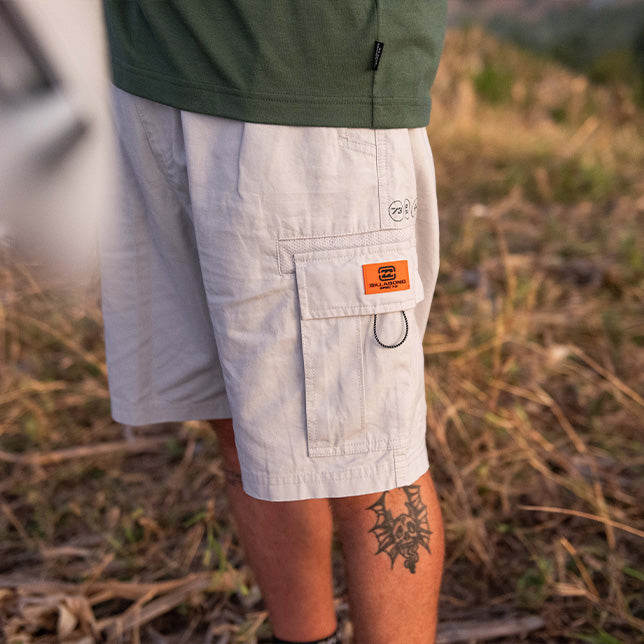 A person stands outdoors in a field, wearing light beige cargo shorts with a visible tattoo on their leg. The background is grassy, conveying a casual, adventurous mood.
