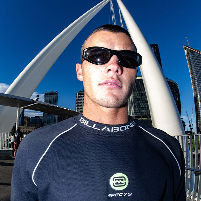 Man in sunglasses and a Billabong jumper stands confidently on a bridge with modern buildings and a clear blue sky in the background. Urban vibe.