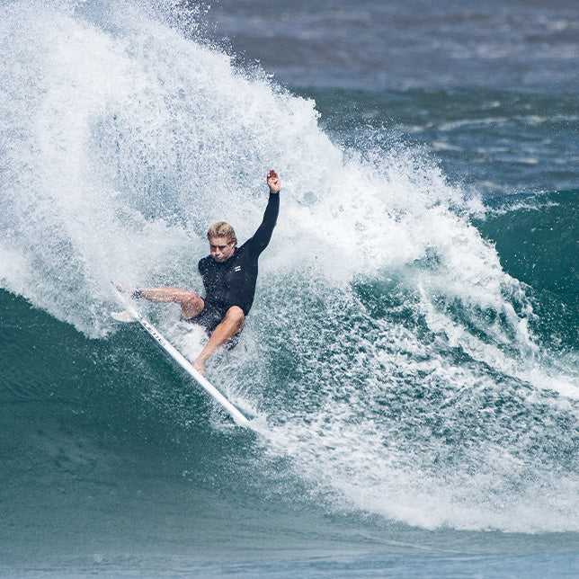 Surfer skillfully does a turn on a crashing wave, water spraying. The surfer is in a wetsuit top and board shorts, capturing surfing in warm water.